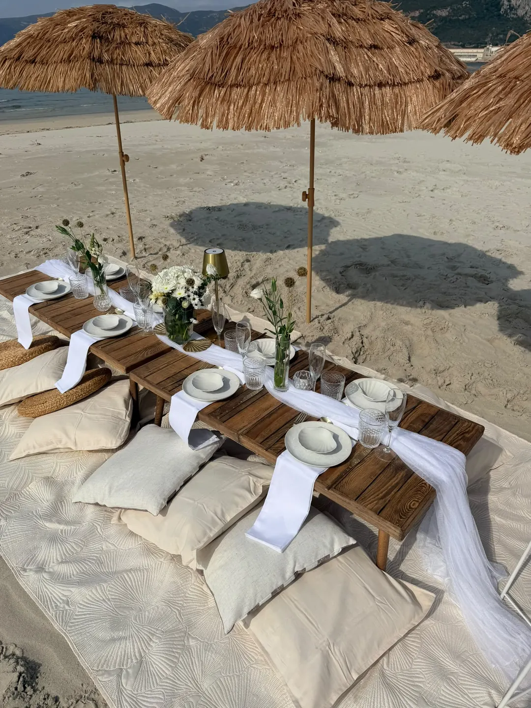 The table laid with white runners, wild flowers and floor cushions, beneath straw parasols