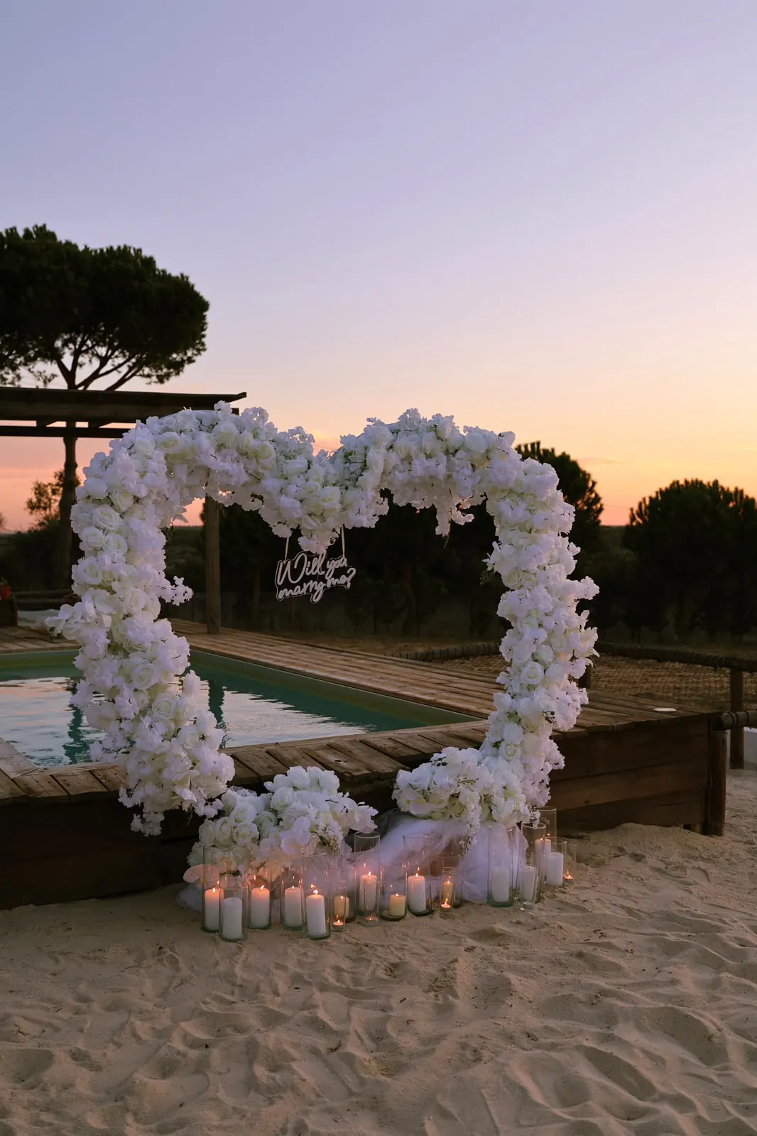 A heart-shaped arch of white flowers by a pool at dusk, with a neon sign reading Will you marry me